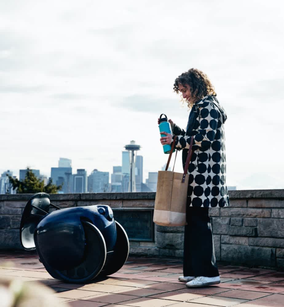 Woman standing on a sidewalk packing items into a twilight blue gita with the Seattle skyline behind her
