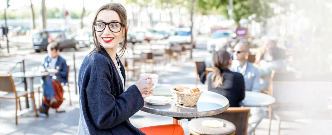 Woman sitting at table outside, holding coffee looking over her shoulder and smiling