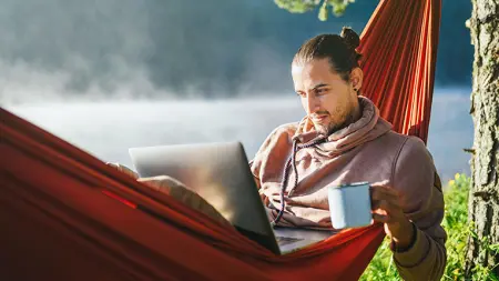 Young man relaxing in an orange hammock by a misty lake holding a cup of coffee, looking at laptop on his lap. 