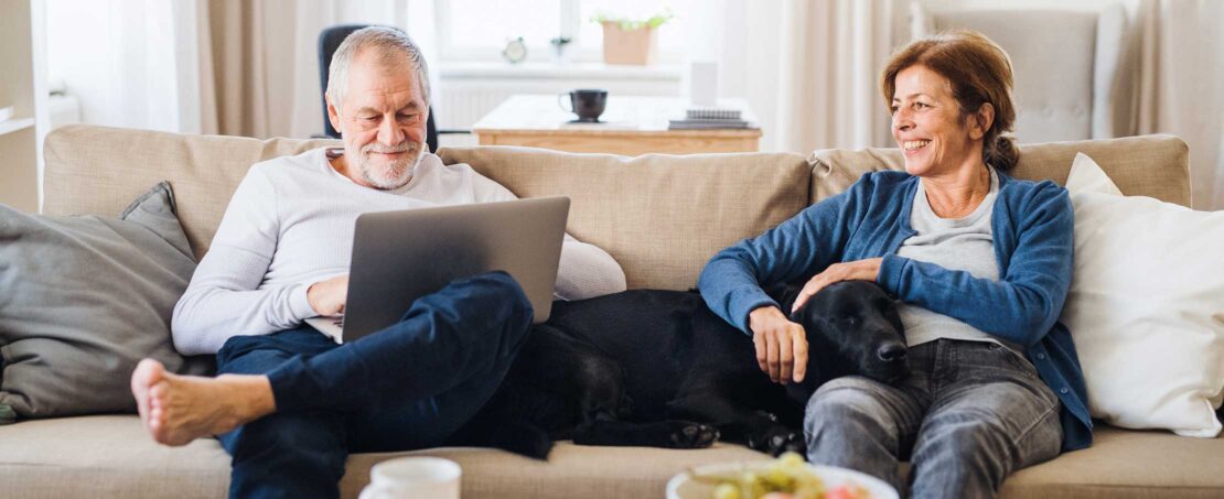 middle aged man with laptop sitting on white couch with woman smiling holding dog