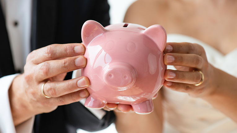 Man and woman wearing wedding attire and wedding rings holding pink piggy bank together