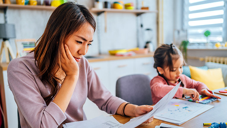 Concerned mother sitting at kitchen table looking at bills while young child sits next to her coloring 