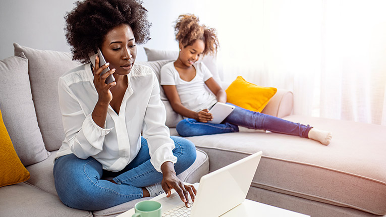 Mother and daughter sitting on couch while mom is talking on the phone and working on laptop.