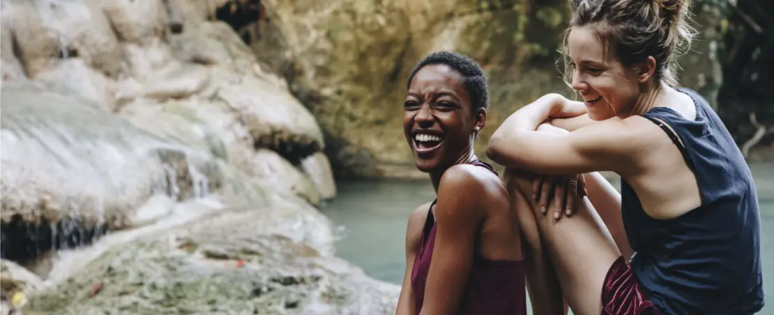 two women smiling sitting on rock near waterfall