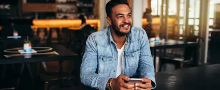 Man in denim button up holding a phone, sitting at table smiling