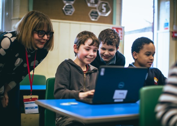 A group of smiling kids and a volunteer cluster around a laptop at a Code Club session.