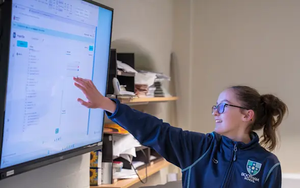 A learner points at her code in the Code Editor interface on a large display in a classroom.