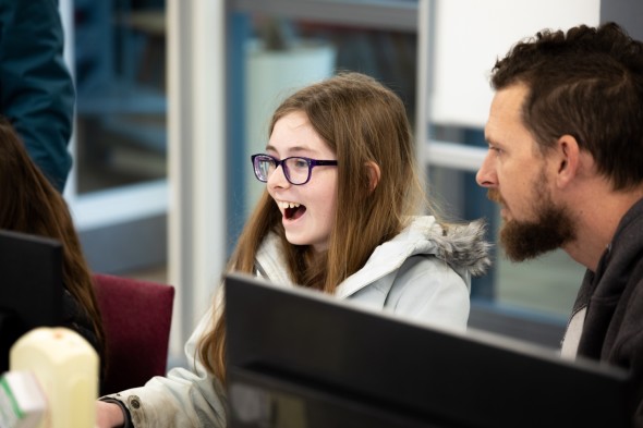 An excited learner gasps at a computer screen while an educator looks on.