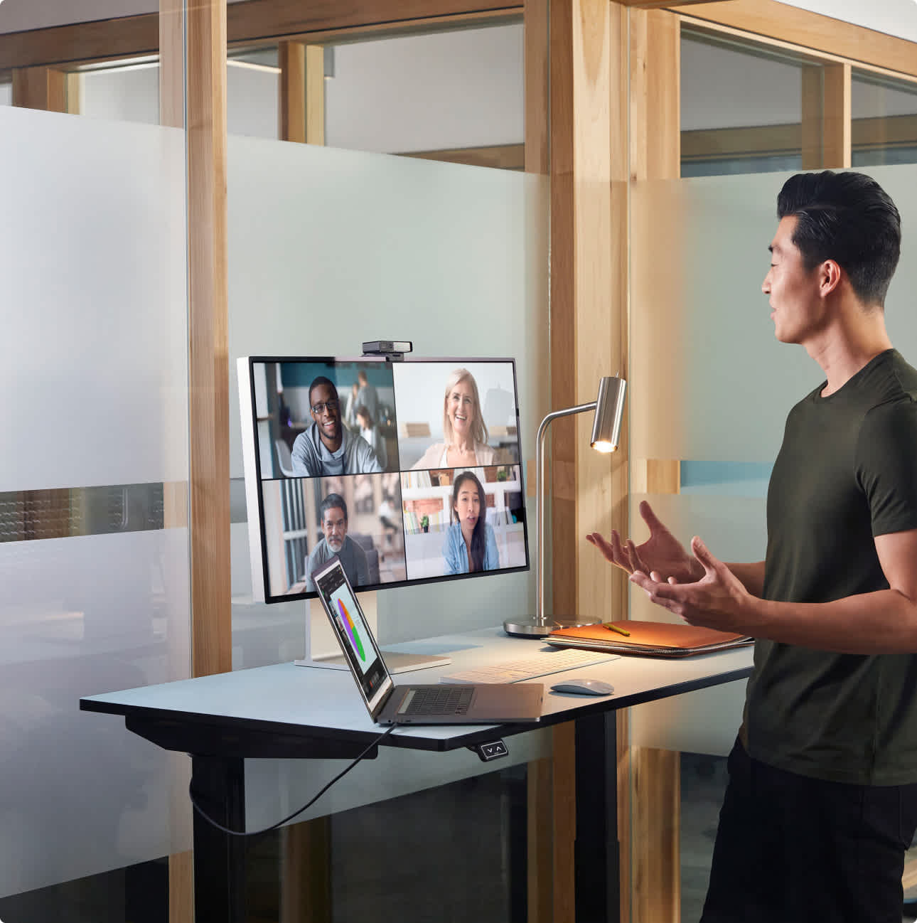 Man standing in a office cubical on a video call with four people on a screen with a Webex Desk Camera attached.