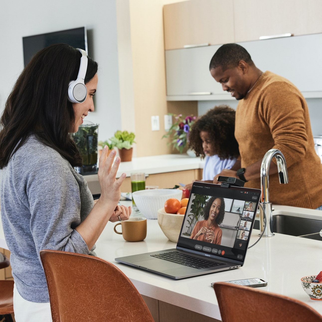 Woman in a video call on her laptop in the kitchen with her family members in the background making food