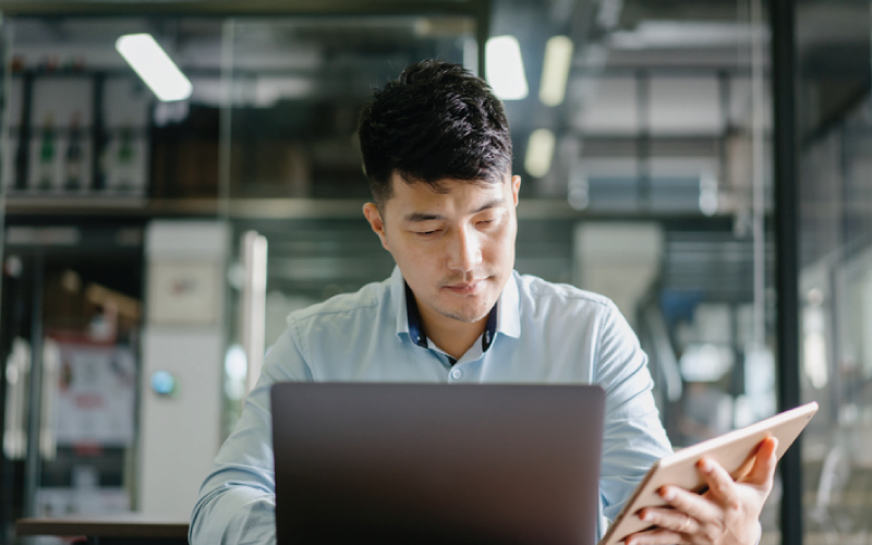 A male employee is reviewing work information on a laptop and tablet