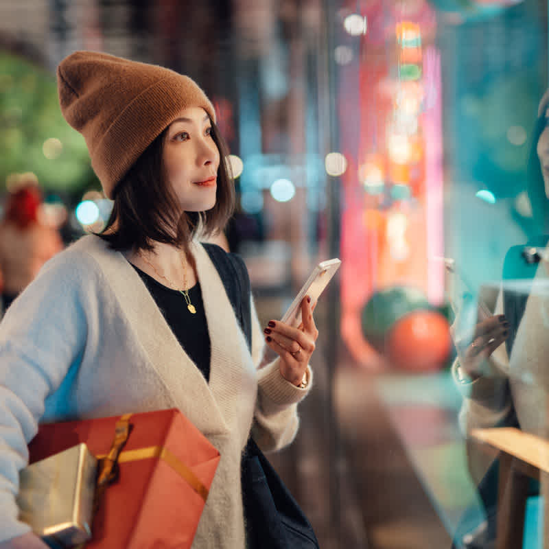 A woman is outside window shopping with her smartphone.