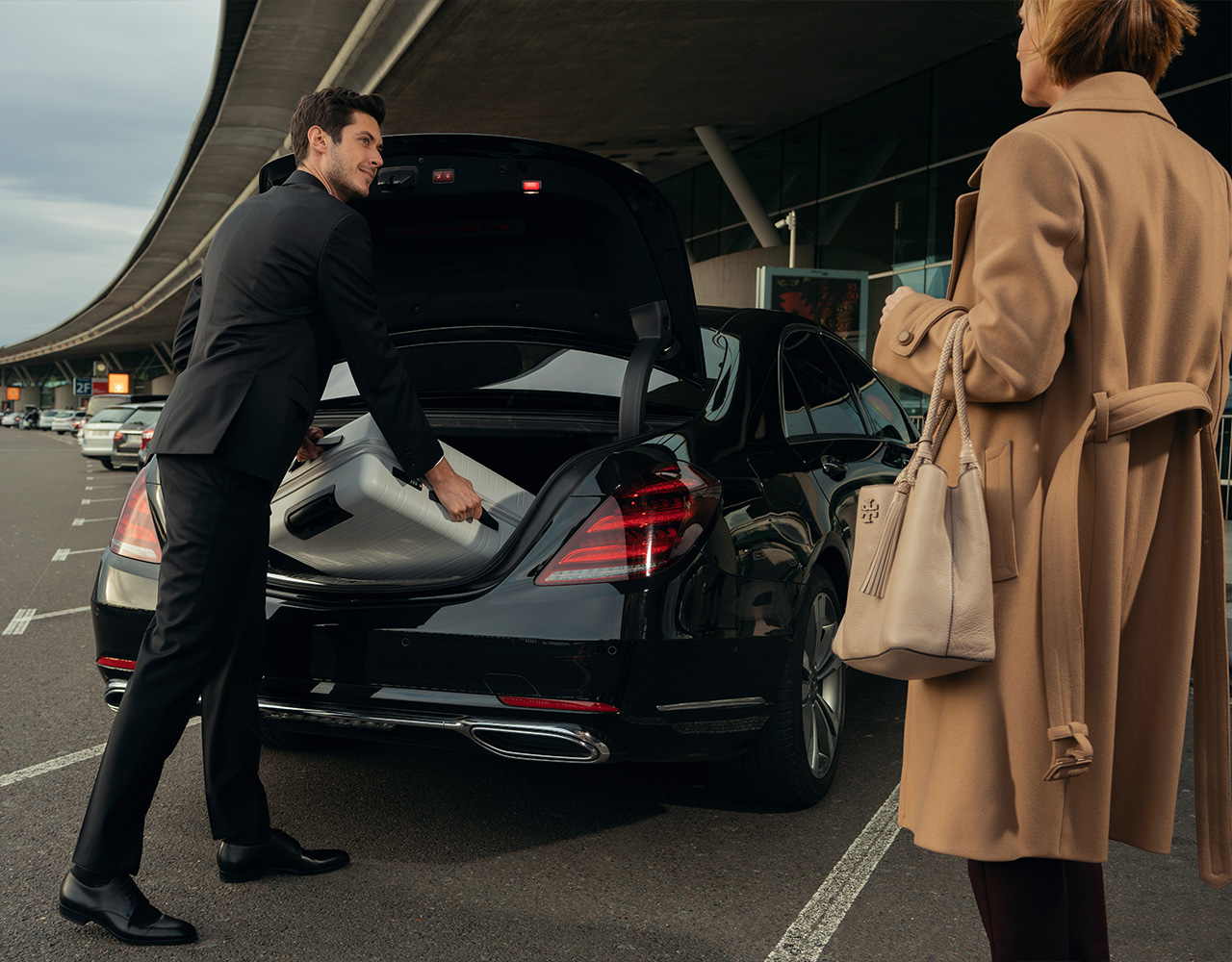 A Blacklane chauffeur in a black suit and tie waits outside arrivals with a tablet pickup sign saying Mr. Doe.