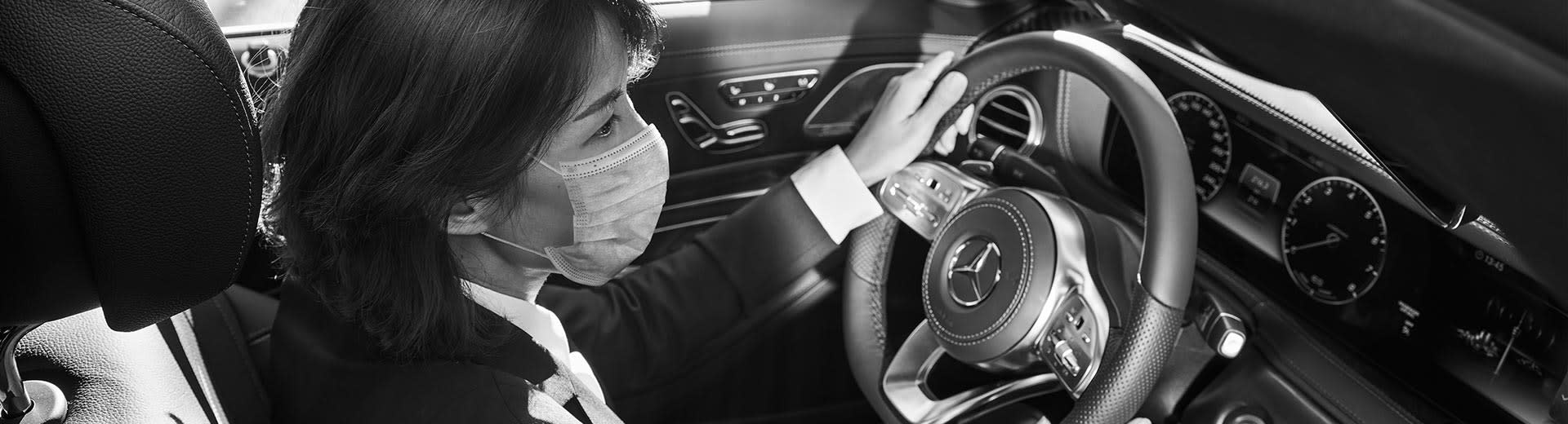 Real Blacklane chauffeur Yen sits behind the wheel of a Mercedes while wearing a face mask.