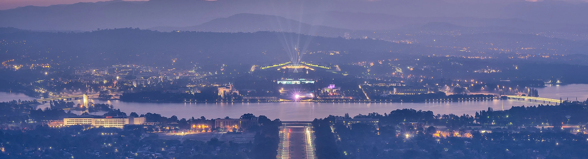 An aerial photograph of Canberra at dusk, with the city sprawled before some mountains and a powerful spotlight piercing the sky.