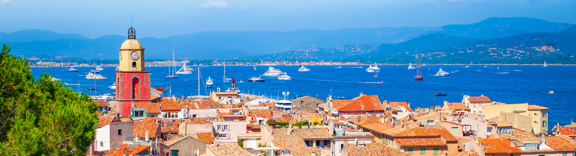 Una vista de puerto de Saint Tropez con un campanario de iglesia y techos en primer plano y agua azul salpicada de botes en el fondo.