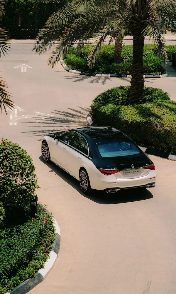 A two-tone Blacklane Mercedes EQS car drives around a roundabout with a palm tree in the background.