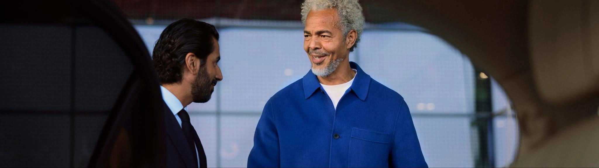 A traveller with grey hair greets his chauffeur while getting into a car outside an airport.