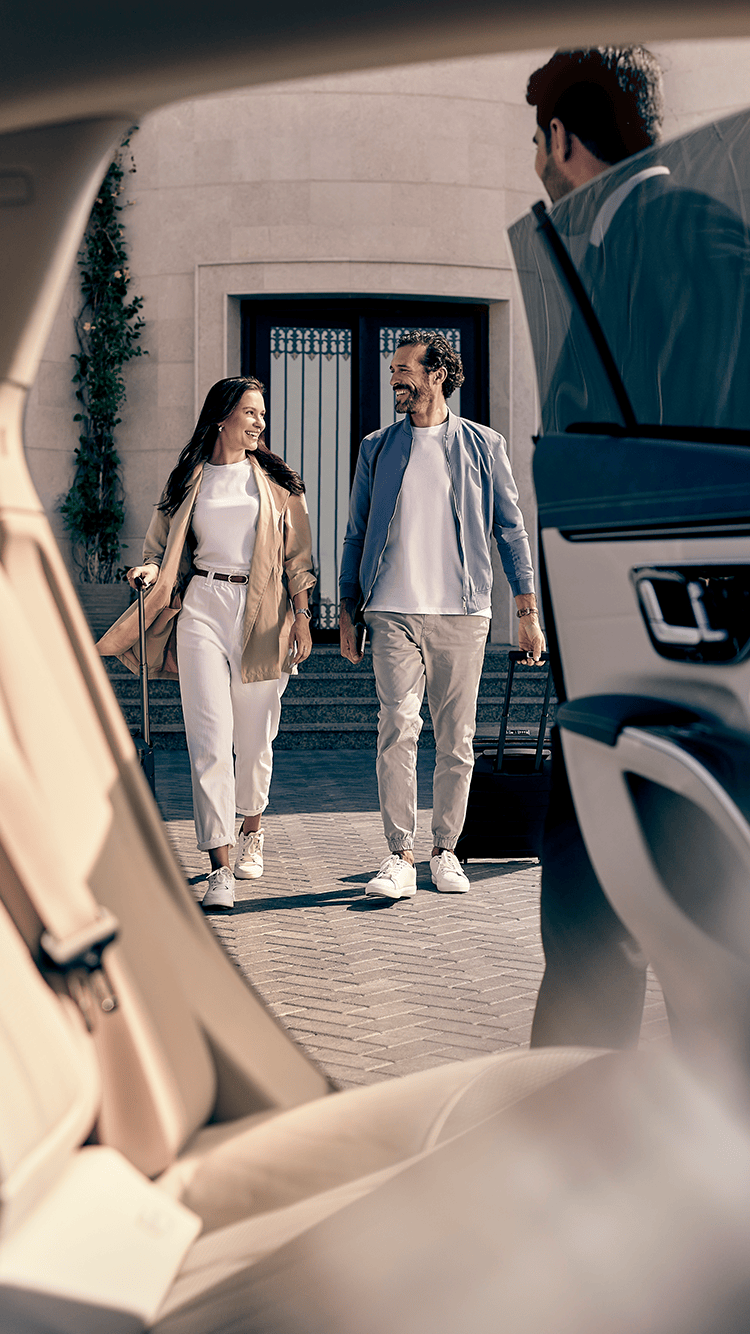 A happy man and woman approach a car, a chauffeur opens the door.