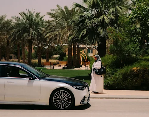 A Mercedes two-tone EQE car with a woman in a headscarf walking towards it with palm trees in the background.