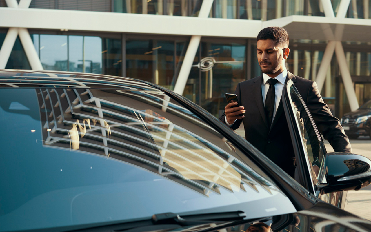A man in a suit stands outside a Mercedes-Benz, looking at his phone.