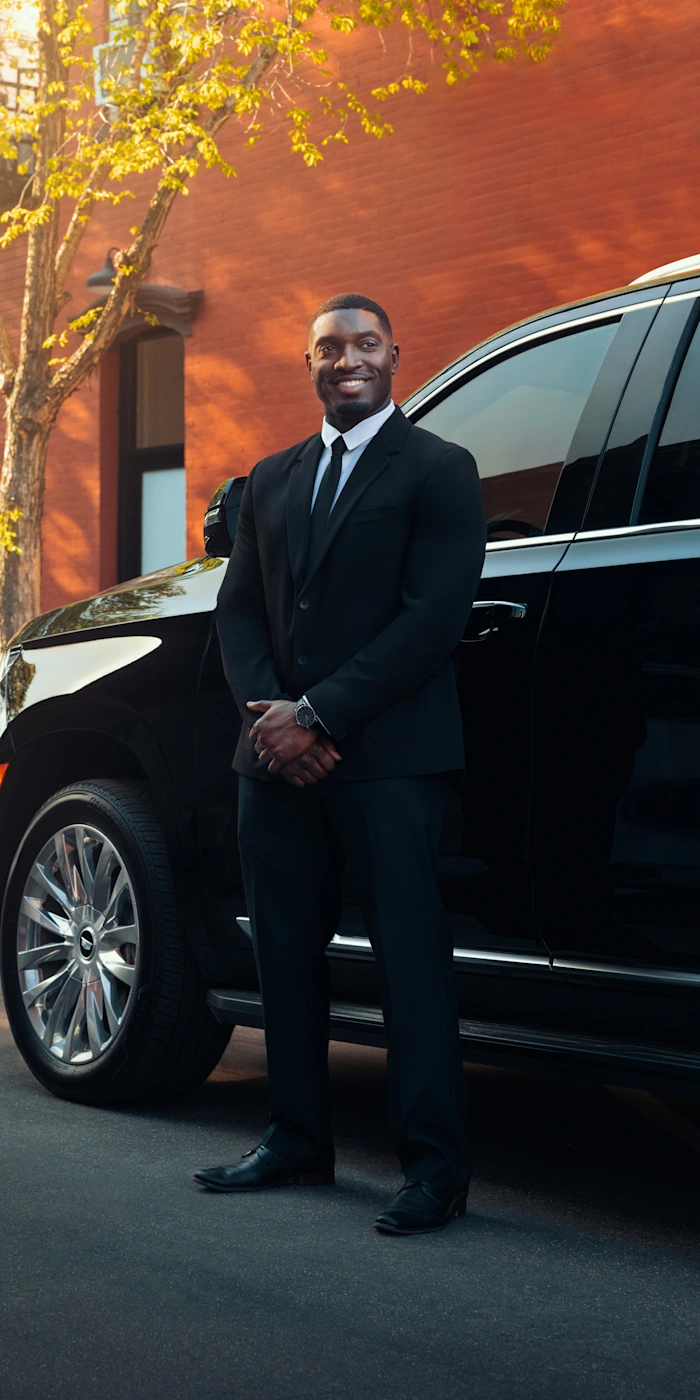 A smiling Blacklane chauffeur stands outside a Business Van limo with a tree and brick wall in the background.