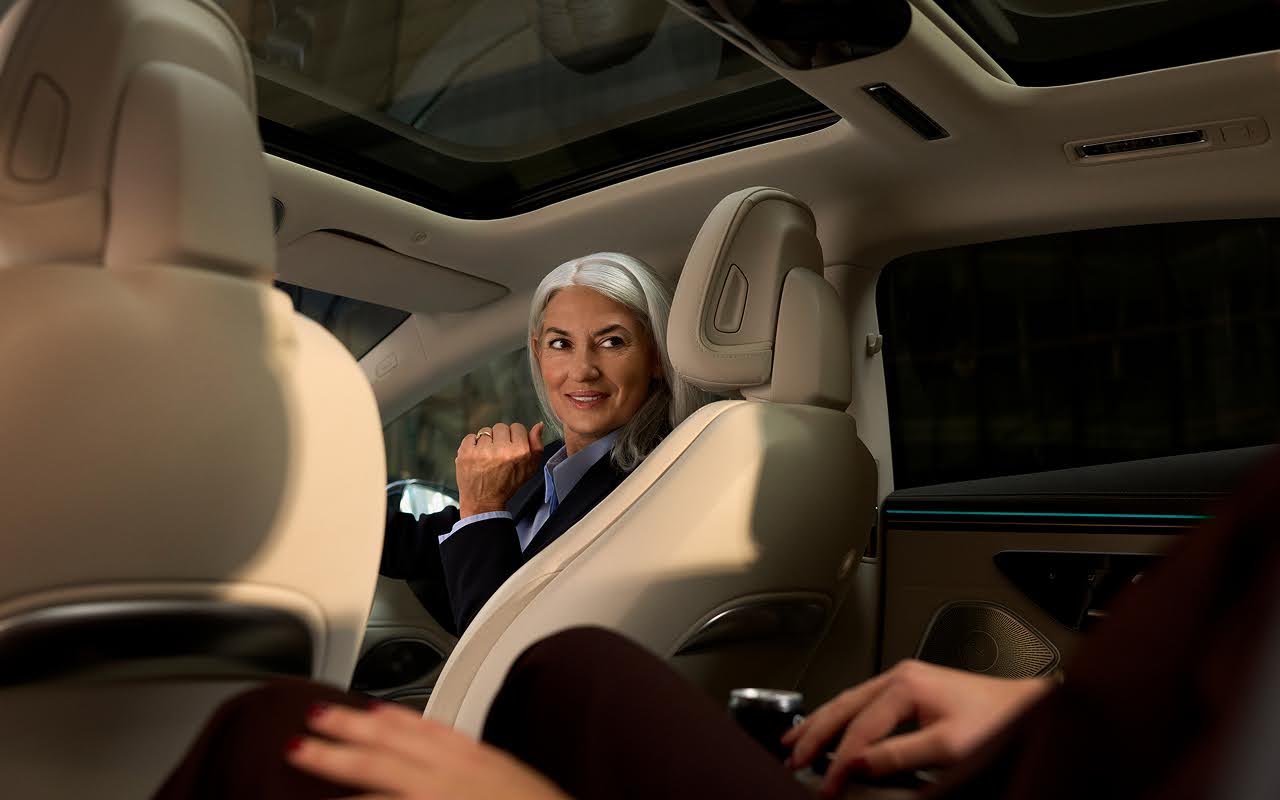 A Blacklane chauffeuse with grey hair turns around in the driver's seat to smile at her passenger.