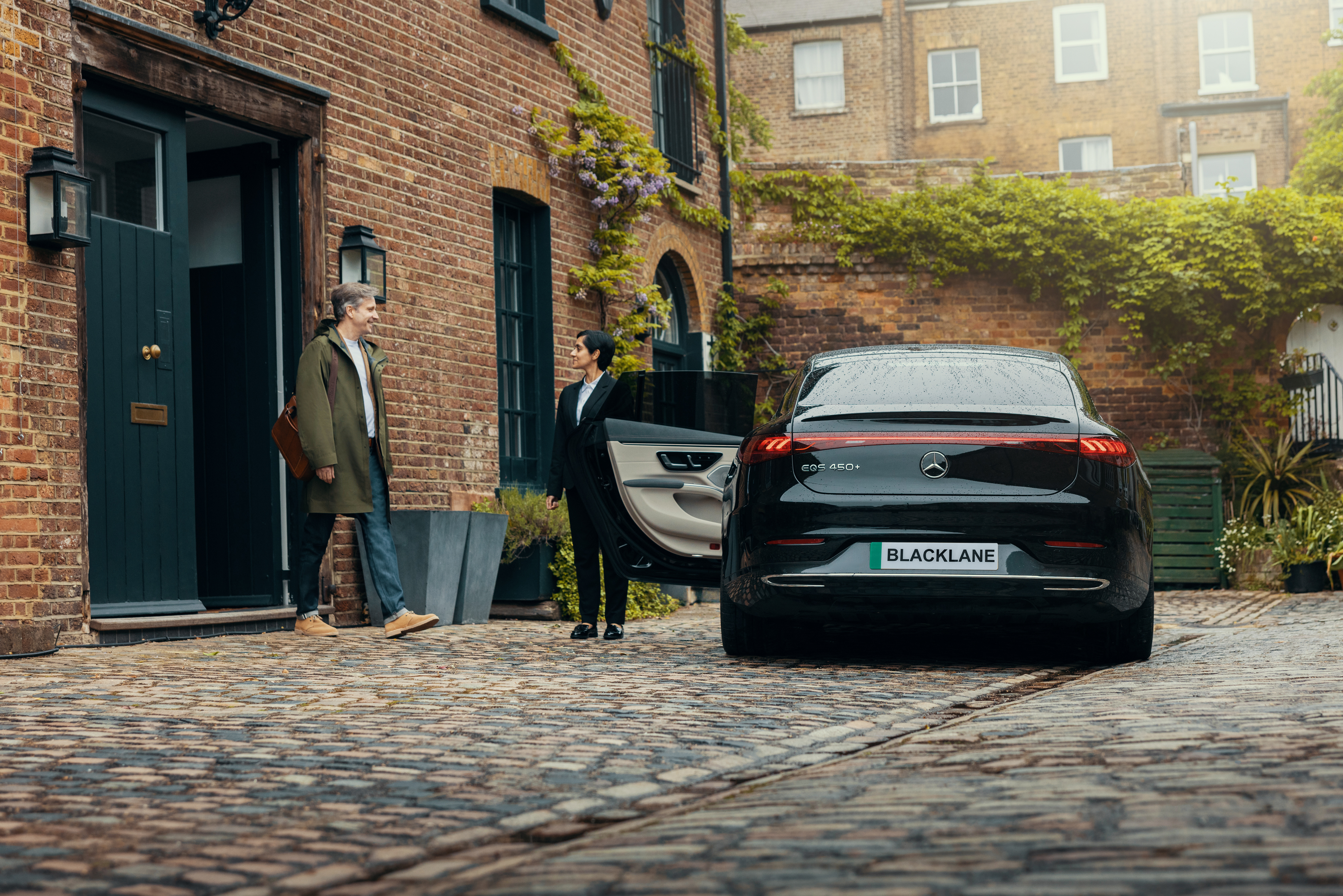 A man leaving his house gets into a Blacklane Mercedes EQS limo while his female chauffeur holds the door open for him.