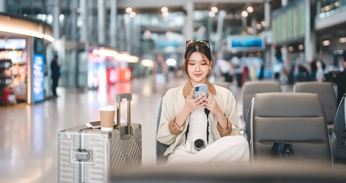 Young asian woman passenger sitting at waiting area international airport terminal using her smartphone app, with her suitcase and coffee beside her.