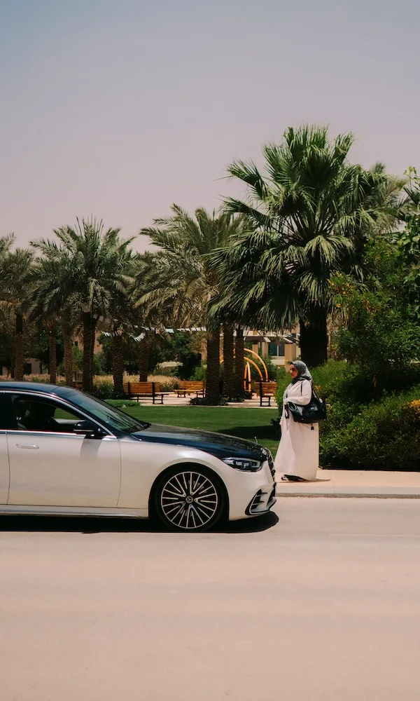 A woman in a headscarf approaches a Blacklane Mercedes EQS limo with palm trees in the background.