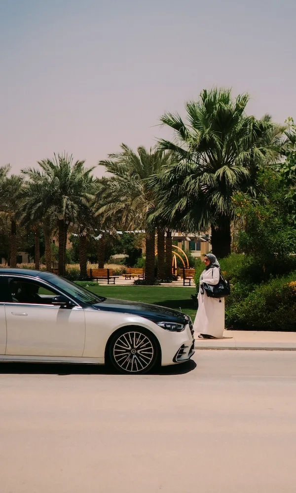 A woman in a headscarf approaches a Blacklane Mercedes EQS limo with palm trees in the background.