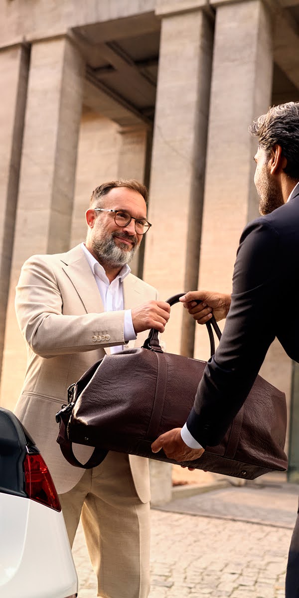 A businessman in a tan suit gets handed his travel back by a suited chauffeur outside a hotel.