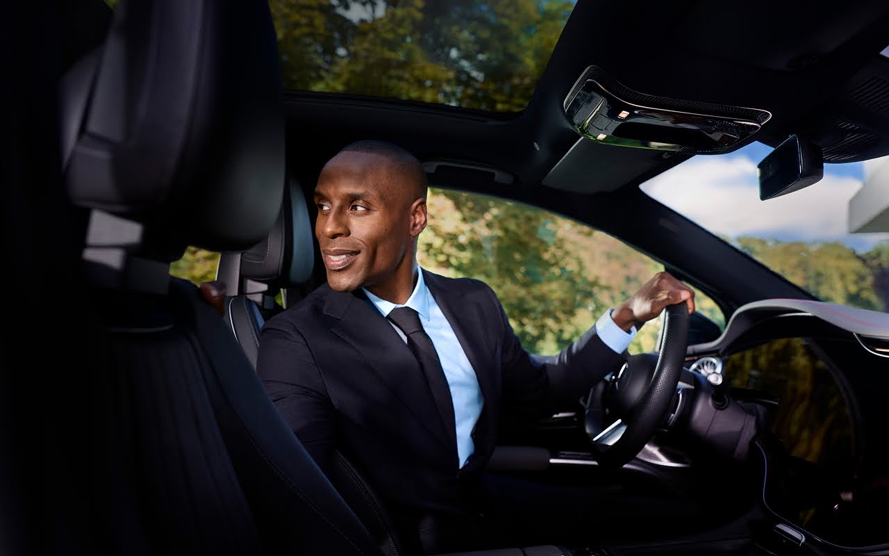 A Blacklane chauffeur sits in the driver's seat of a limousine, smiling and looking over his should into the back seat.