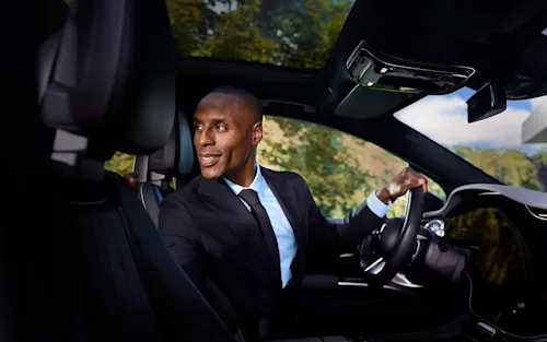 A Blacklane chauffeur sits in the driver's seat of a limousine, smiling and looking over his should into the back seat.