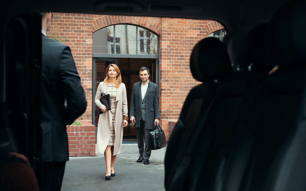 A well-dressed couple approaches their Sprinter Van with their chauffeur standing by.