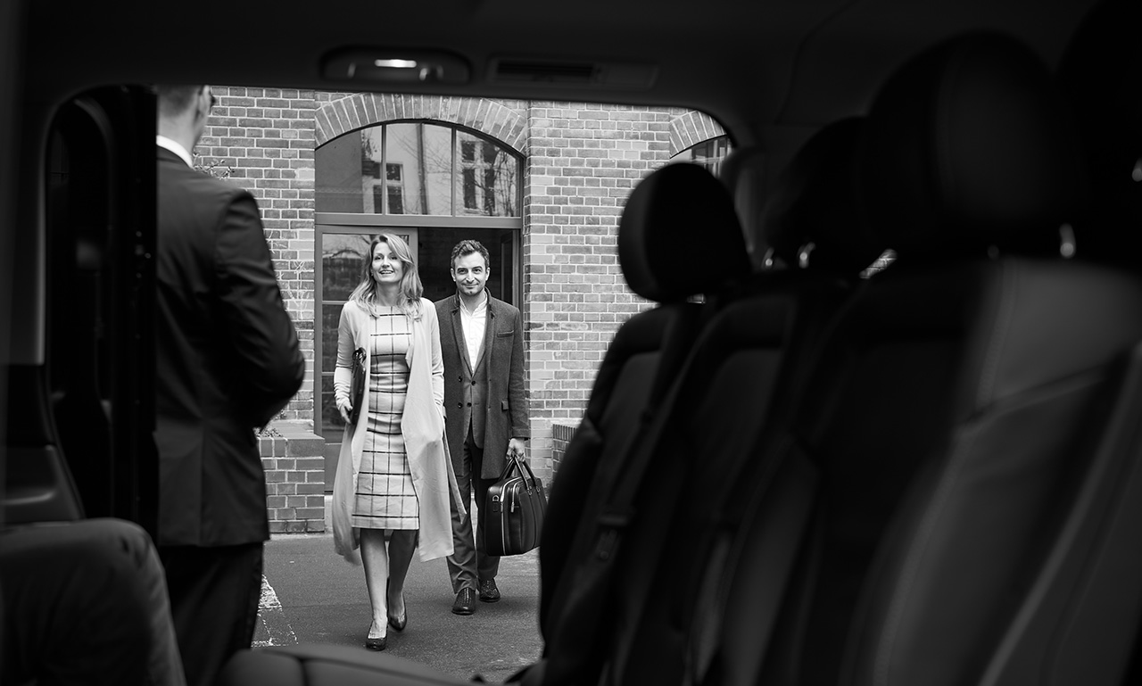 A well-dressed couple approaches their Sprinter Van with their chauffeur standing by.