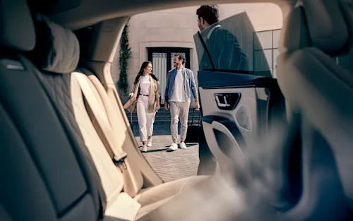 A couple with rolling luggage walk toward their chauffeur who holds the door of a Mercedes-Benz for them.