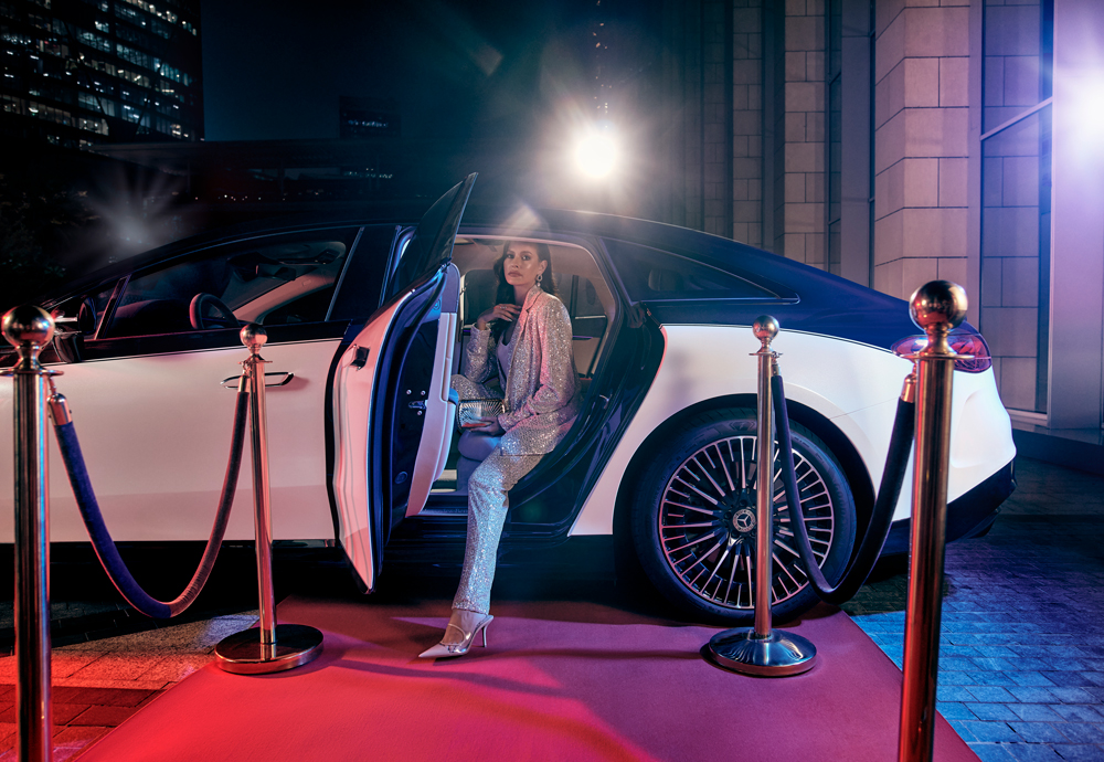 Una glamurosa pasajera sale por la puerta trasera de un Mercedes EQS bicolor hacia una alfombra roja.