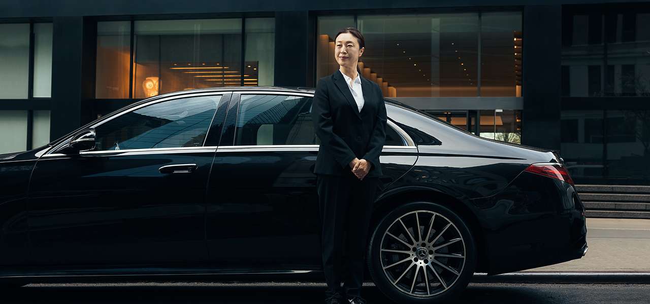 A female chauffeur in a suit stands beside a Blacklane Mercedes limo.