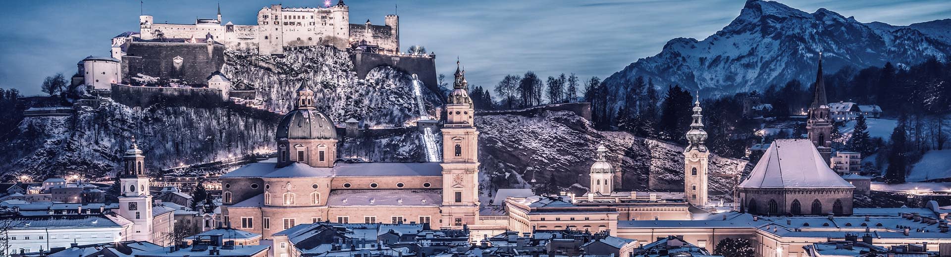 El castillo de Salzburgo ilumina el cielo nocturno en el fondo, mientras que los edificios históricos son visibles en primer plano.
