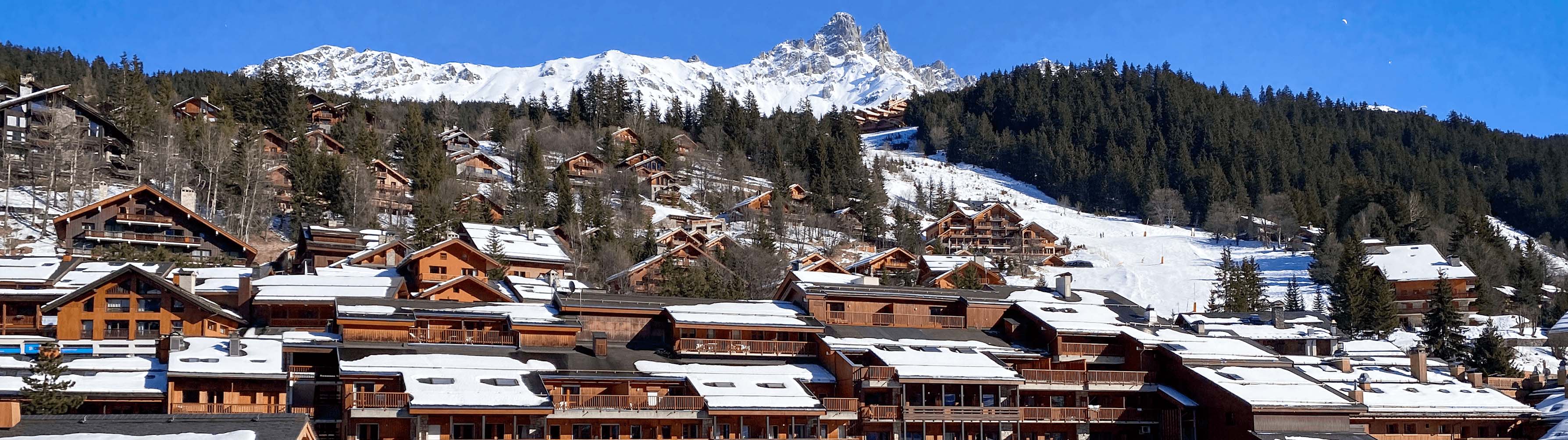 Brown chalets in Meribel stand before snow-packed slopes on a clear and sunny day.