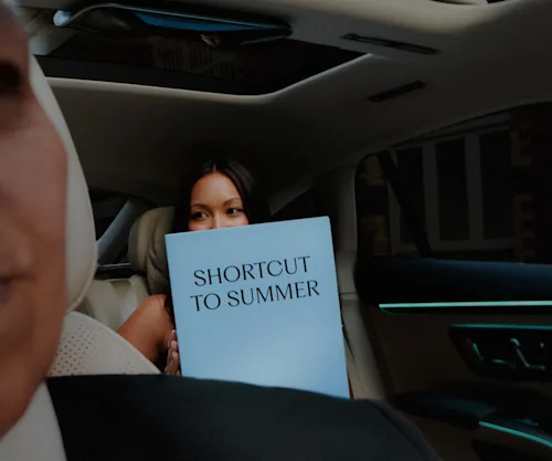A young woman sits in the backseat of a Blacklane limo holding a sign saying "Shortcut to Summer" with her chauffeur in the left foreground.