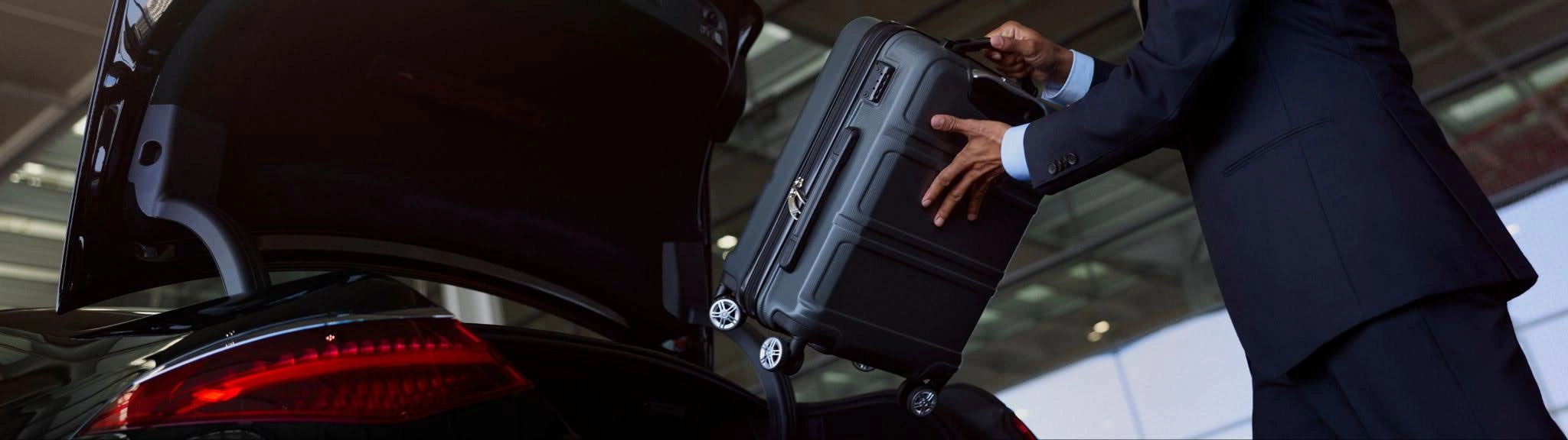 A suited Blacklane chauffeur places a luggage case in the back seat of a black Mercedes luxury vehicle.