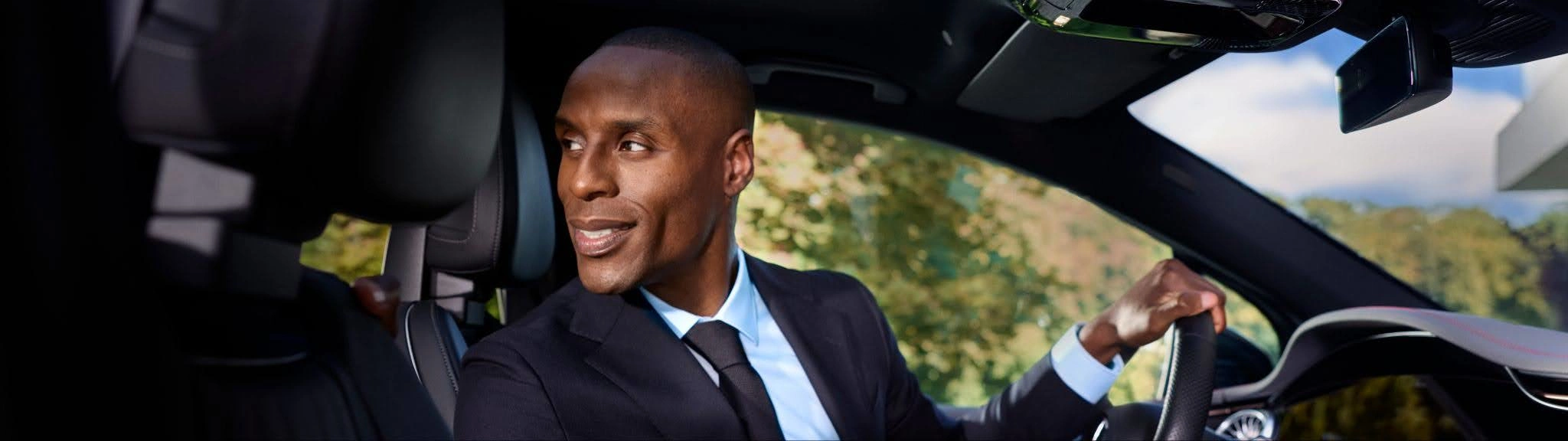 A Blacklane chauffeur sits in the driver's seat of a limousine, smiling and looking over his should into the back seat.