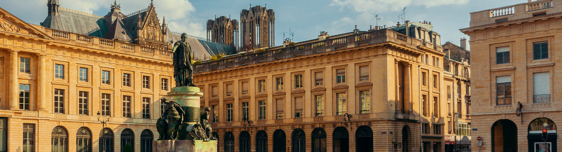 A picture of Place Royale, a square in Reims. In the foreground stands a bronze statue of Louis XV. The twin towers of Cathédrale Notre-Dame are visible above buildings in the background.