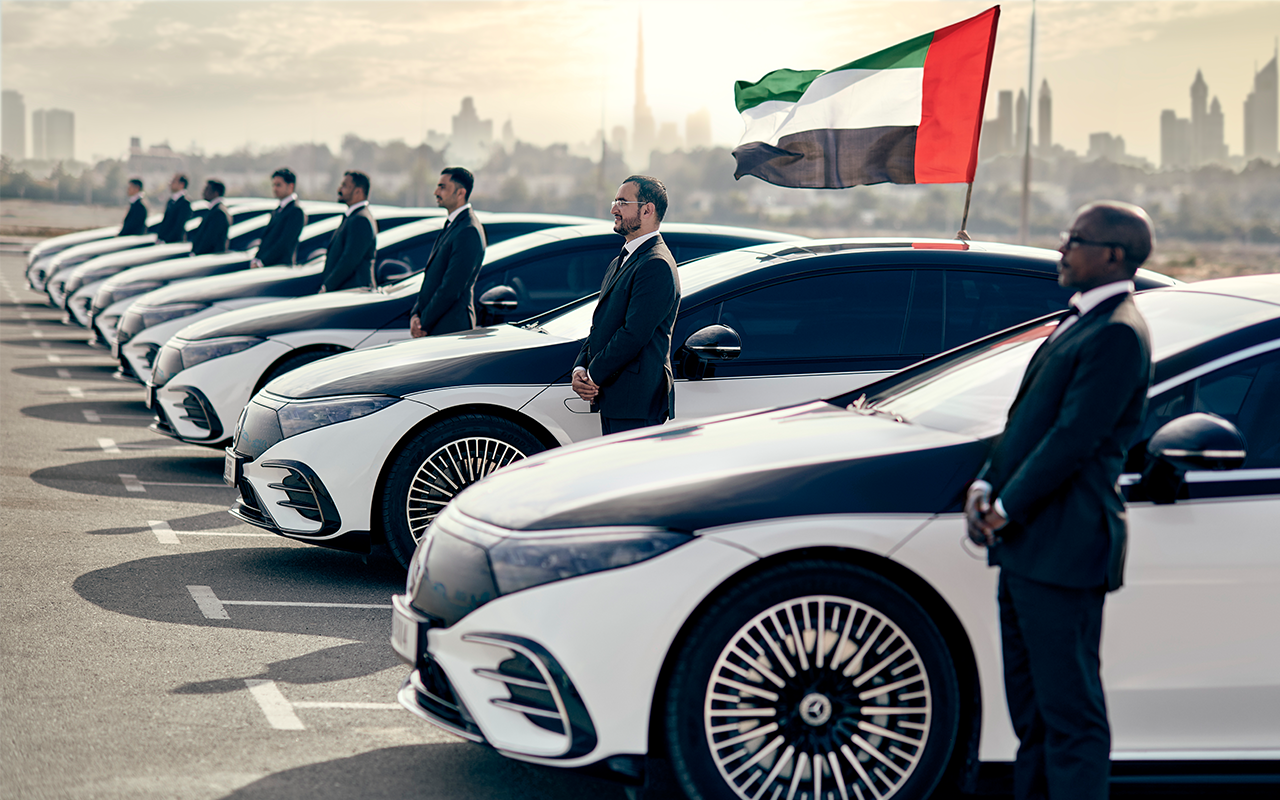 A row of Blacklane chauffeurs standing next to their parked Mercedes-Benz EQSes with the Emirati flag flying behind them.