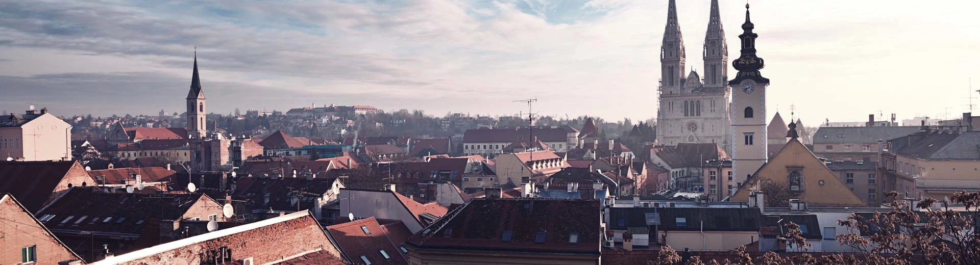 Dächer und Kirchenstätten dominieren die Skyline von Zagreb unter einem grauen Himmel und der späten Sonne.