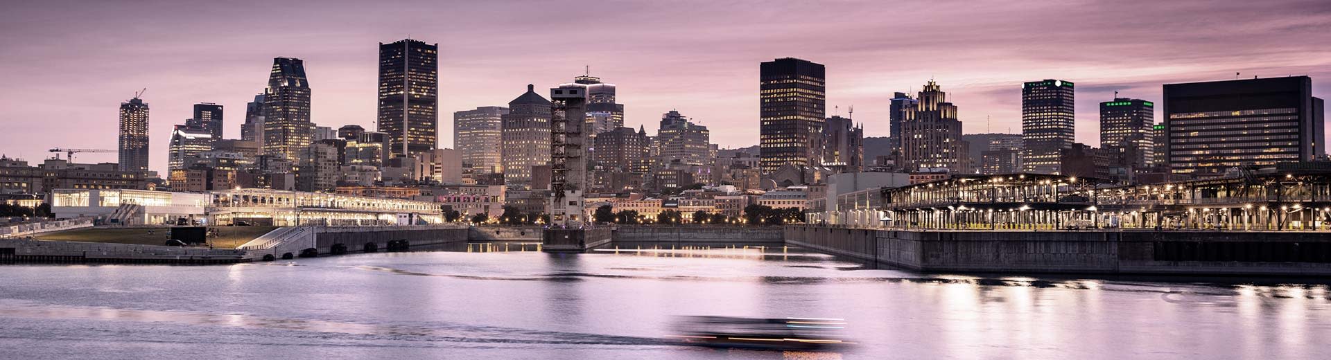 Contre un ciel violet, nous voyons les gratte-ciel du centre-ville de Montréal devant un plan d'eau sombre.		