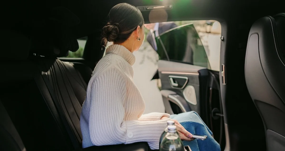 A female passenger in the backseat of a Blacklane holds her phone while looking out the open car door the chauffeur is holding for her.