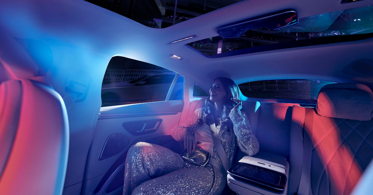 A woman in a sparkling outfit sits in a luxury car's backseat, bathed in blue and pink lights, exuding a glamorous and relaxed vibe at night.